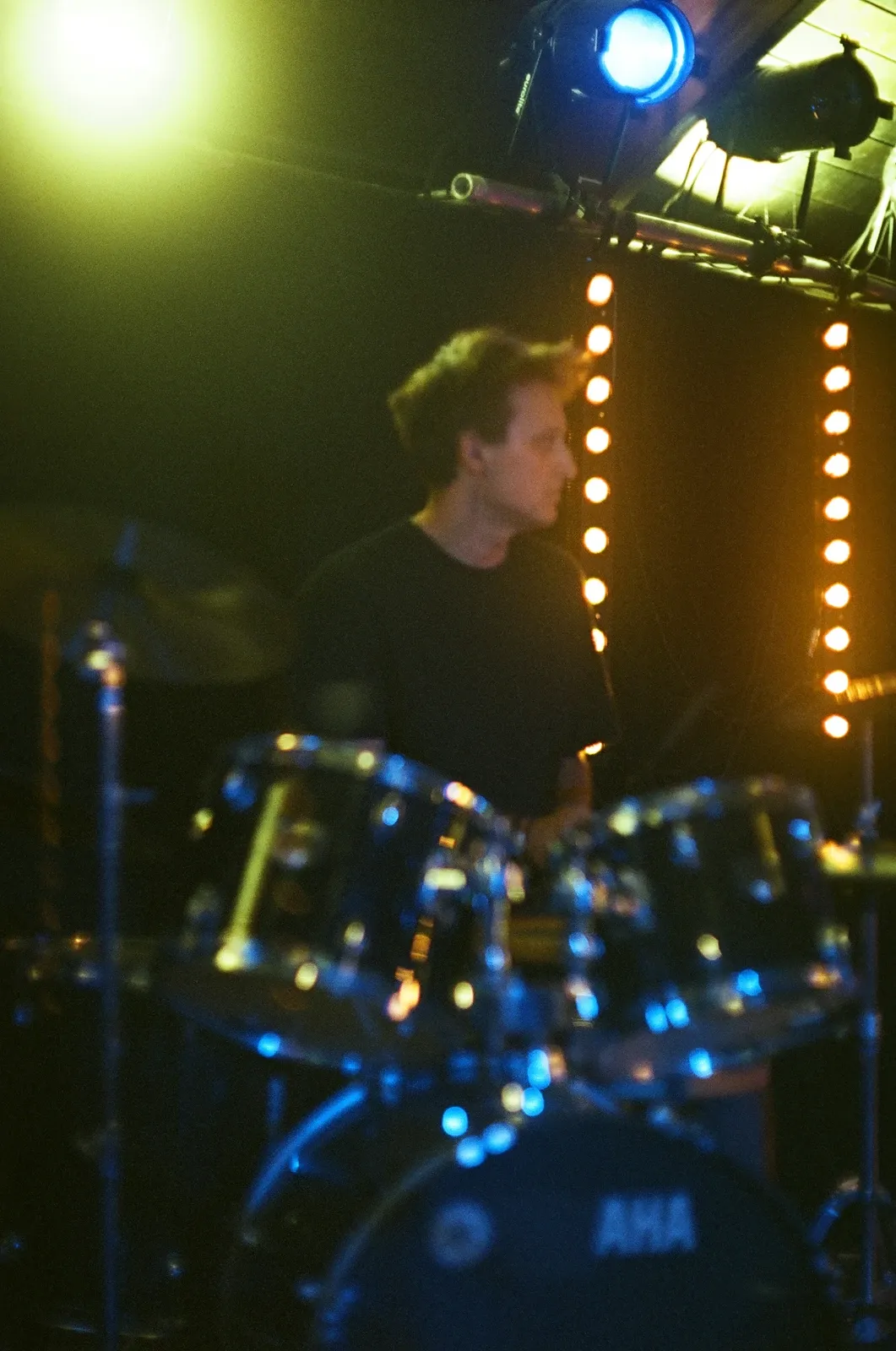 The drummer of Chroustones behind the kit, seen through a cymbal stand under a yellow spotlight